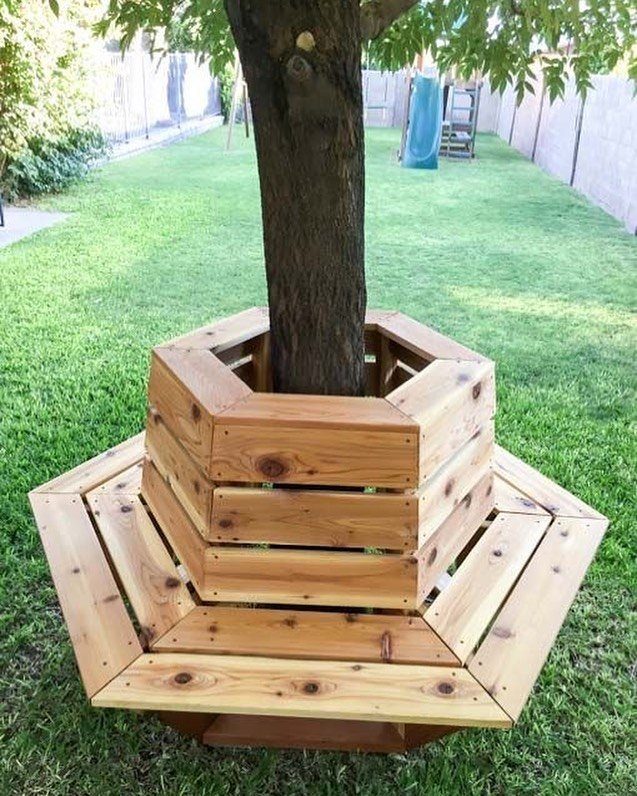 A wooden hexagonal bench encircling the base of a tree trunk on a green lawn.