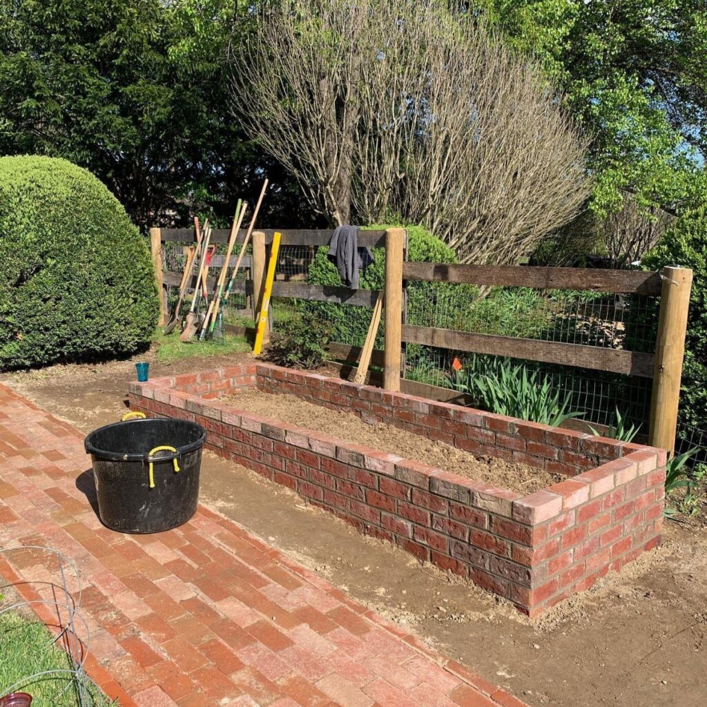 Red brick raised garden bed situated next to a brick walkway in a garden.