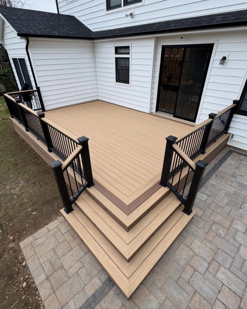 Tan composite deck with black railings and wide steps leading down to a grey stone patio.