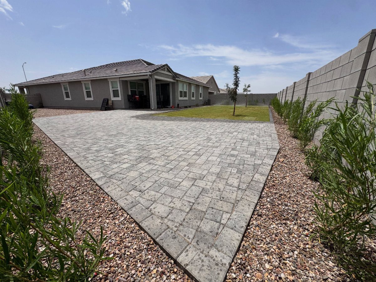 Large gray stone paver patio surrounded by gravel and green shrubs in a backyard.