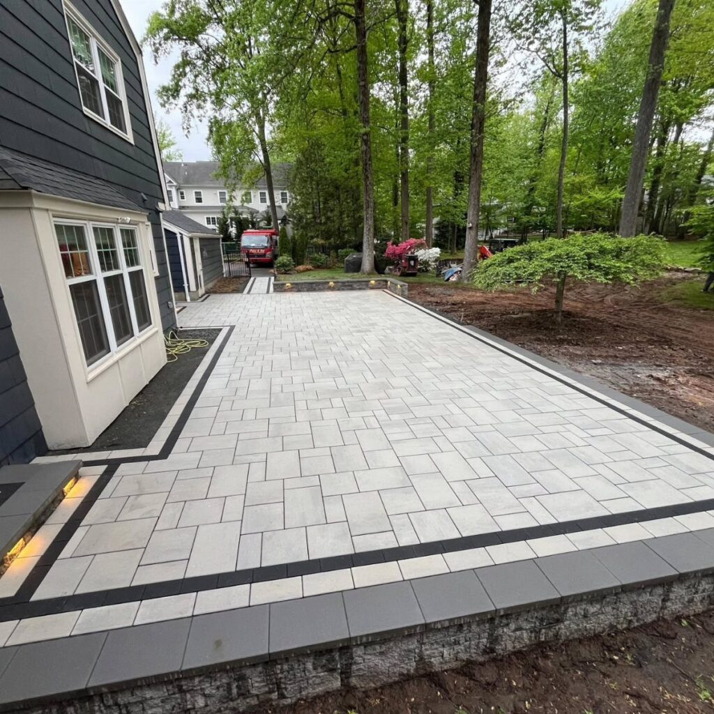 Light gray stone paver patio with dark borders next to a blue house and green trees.