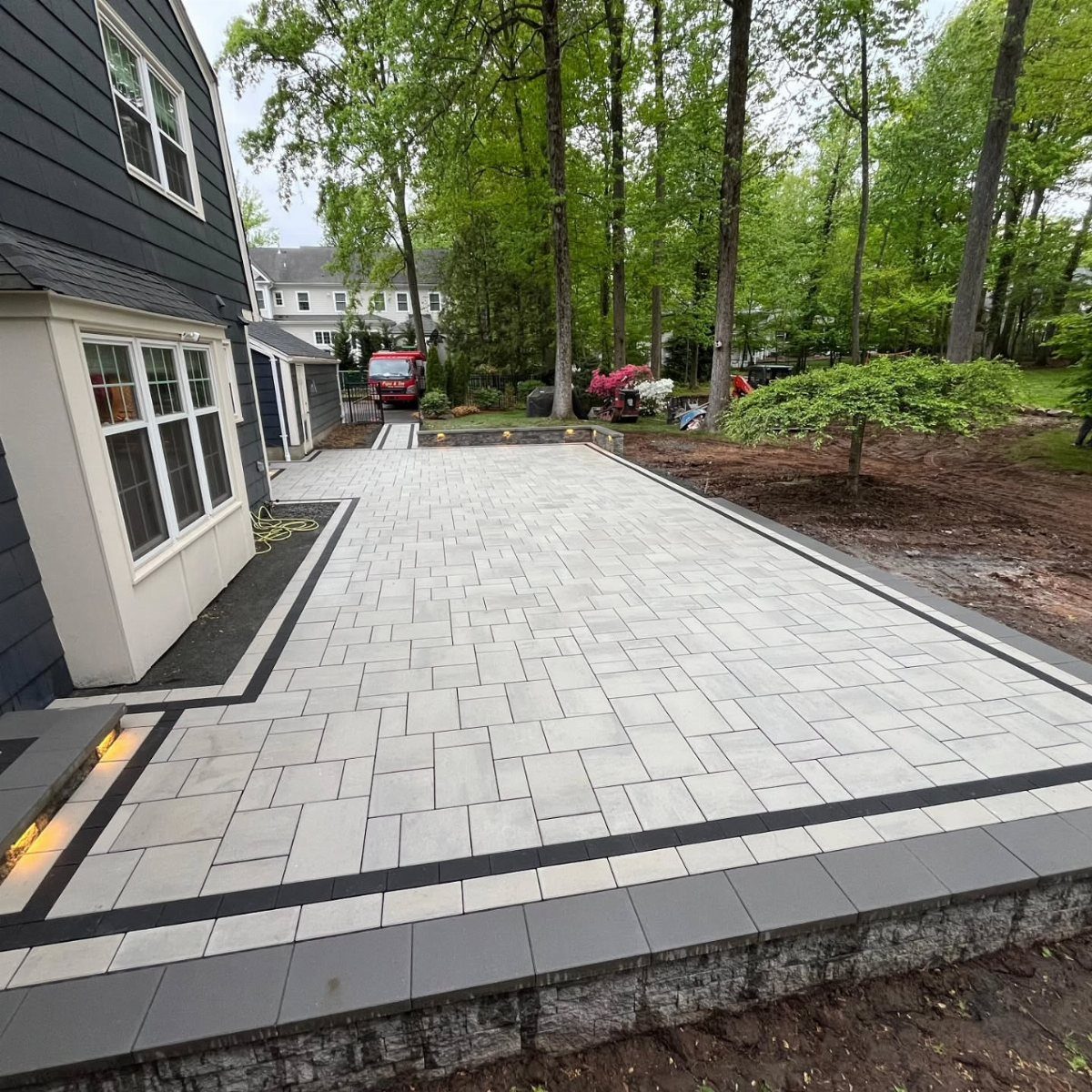 Light gray stone paver patio with dark borders next to a blue house and green trees.