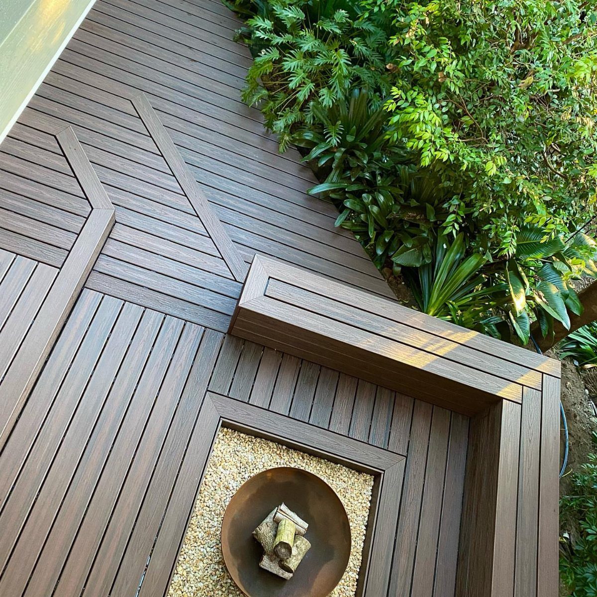 Dark brown composite decking with geometric inlay patterns and a built-in fire pit area surrounded by greenery.