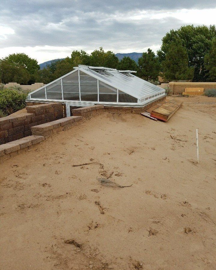 A white-framed glass greenhouse structure being built into a sandy hillside with stone retaining walls.